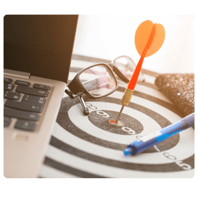 A laptop, pair of glasses, pen, and notebook siting on a target with a dart in the bullseye.