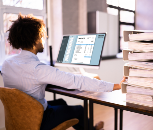 A person working on a desktop computer with a pile of paperfilled binders next to the person.