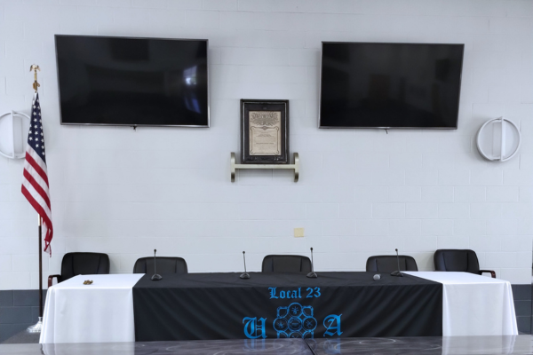 Two video displays on a wall behind a long table.