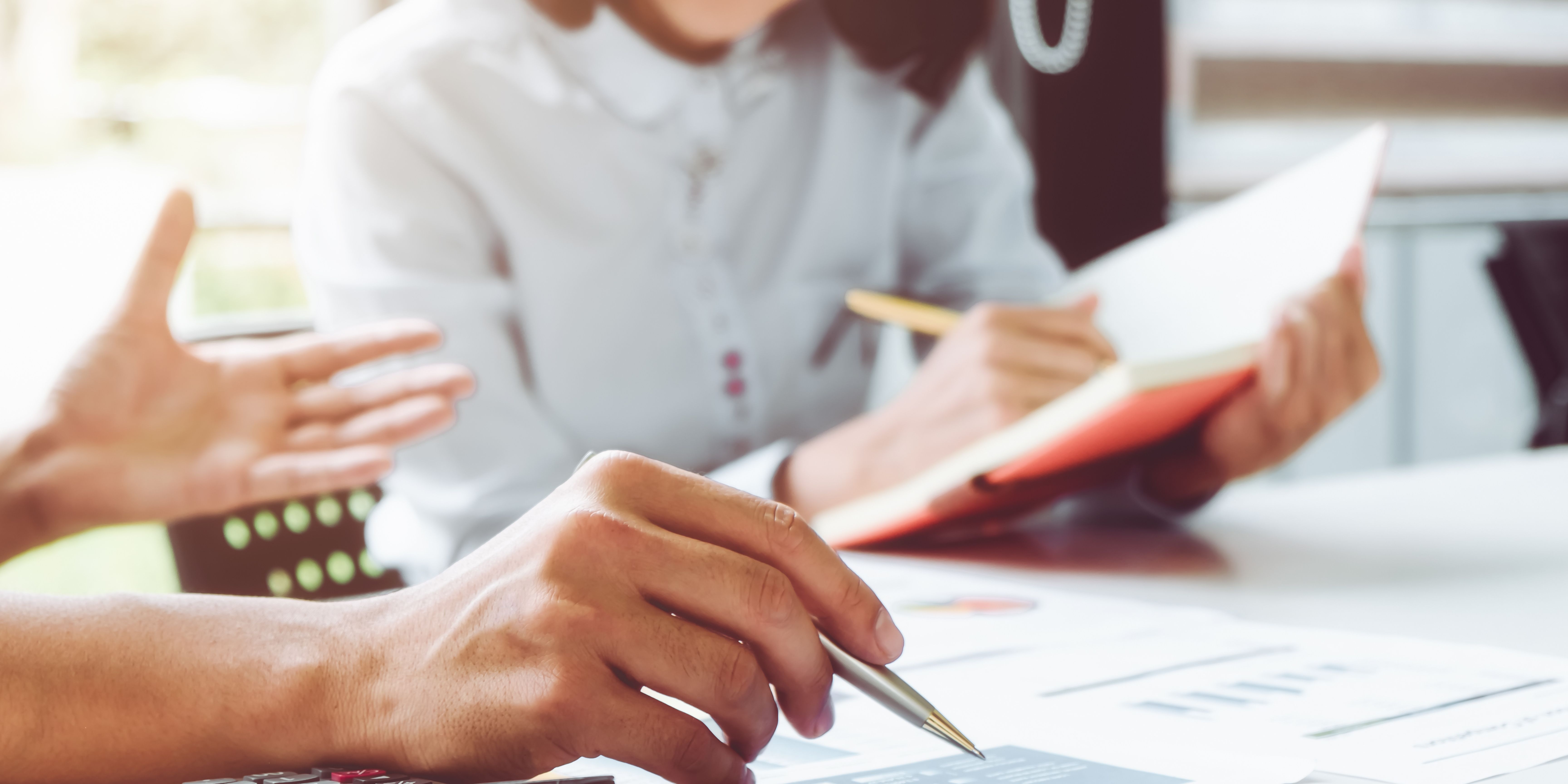 Two people working at a table. One of them is pointing to a piece of paper with their pen.