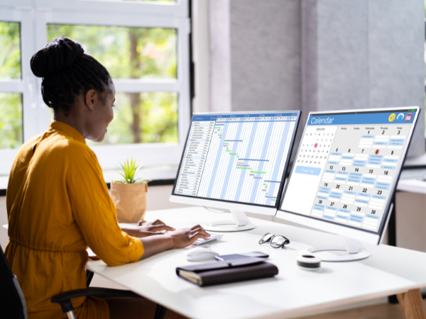 A person with two computer monitors showing a calendar and scheduling on them.