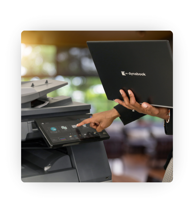 An image of a person holding a dynabook laptop and using a Sharp multifunctional printer.